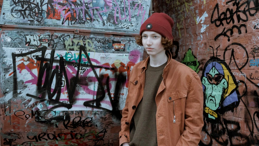 a man standing in front of a wall covered in graffiti, an album cover, pexels, red haired teen boy, beanie, very very very pale skin, caracter with brown hat