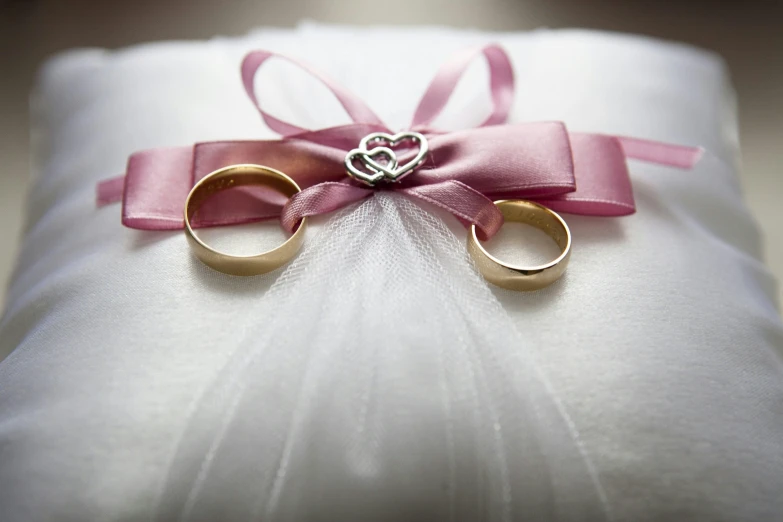 a couple of wedding rings sitting on top of a pillow, by Sylvia Wishart, pexels, ribbon, white and pink, hearts, actual photo