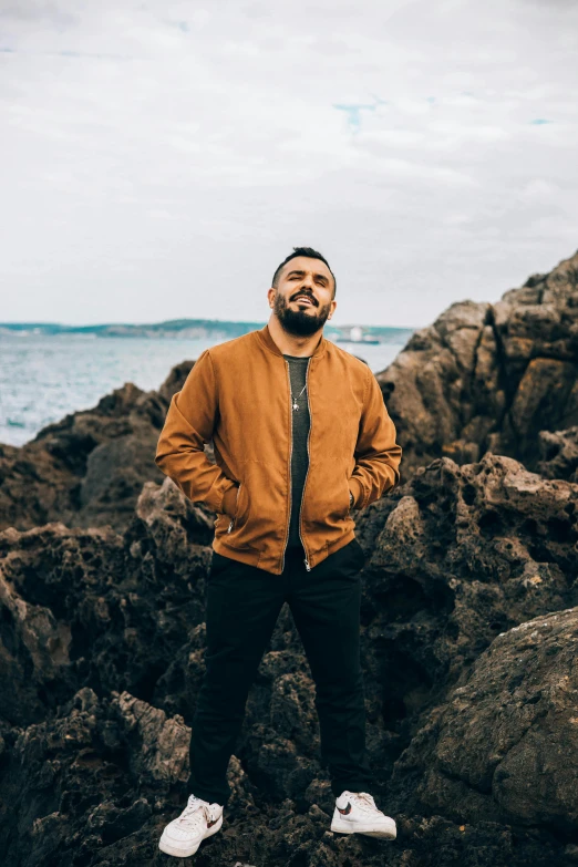 a man standing on a rocky beach next to the ocean, a portrait of rahul kohli, he is wearing a brown sweater, standing in gold foil, wearing a turtleneck and jacket