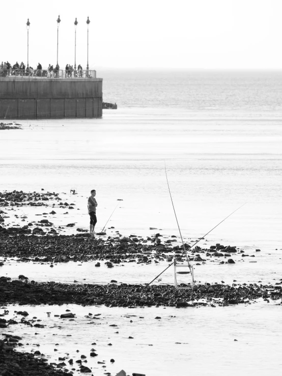 a man standing on top of a beach next to a body of water, a black and white photo, by Tony Tuckson, people angling at the edge, harbour, by greg rutkowski, spectators
