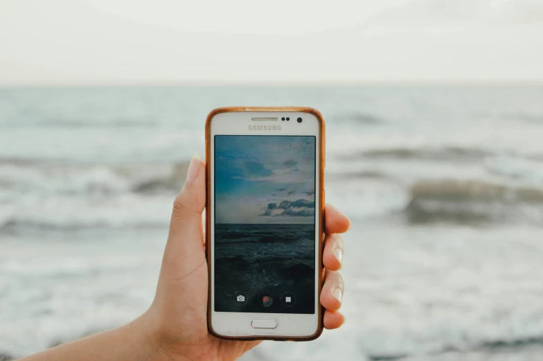 a person taking a picture of the ocean with their cell phone, pexels, holding a galaxy, slightly golden, android format, looking directly at the viewer