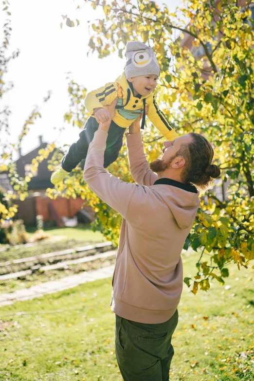 a man holding a baby up in the air, in the garden, wearing a yellow hoodie, canvas, thumbnail