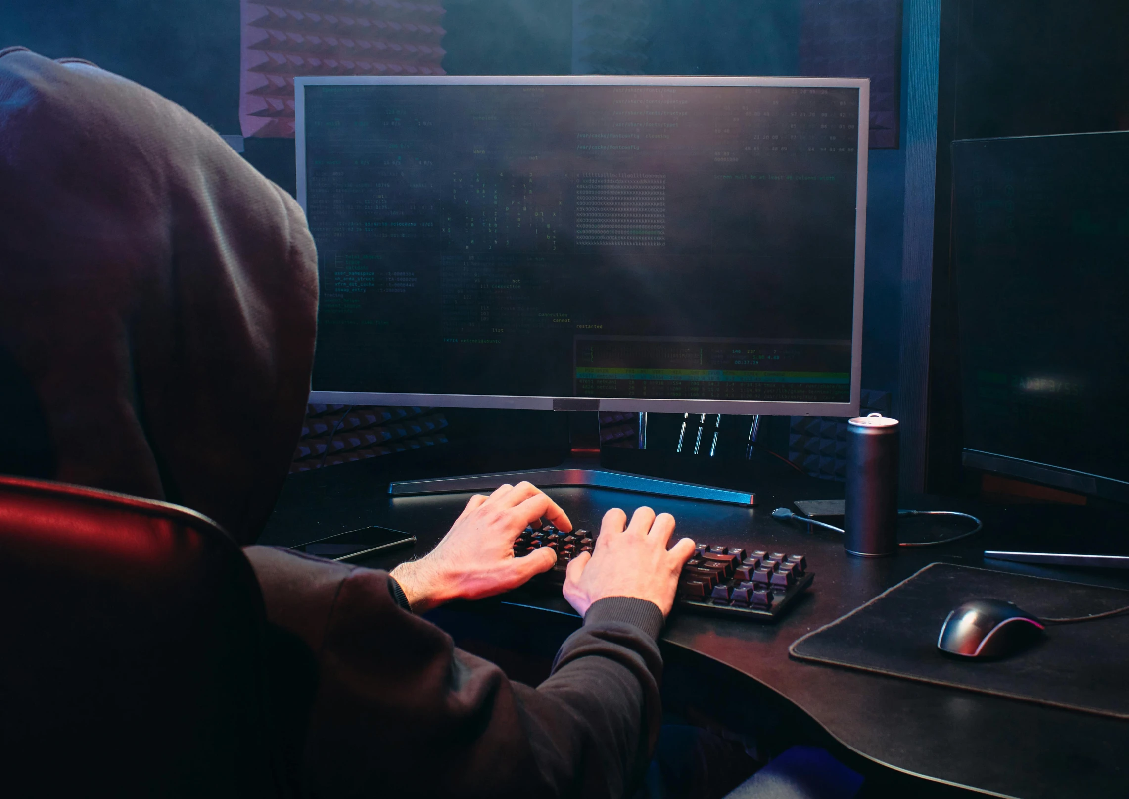 a person sitting at a desk in front of a computer, wearing a dark hood, cryptoblades, console and computer, background image