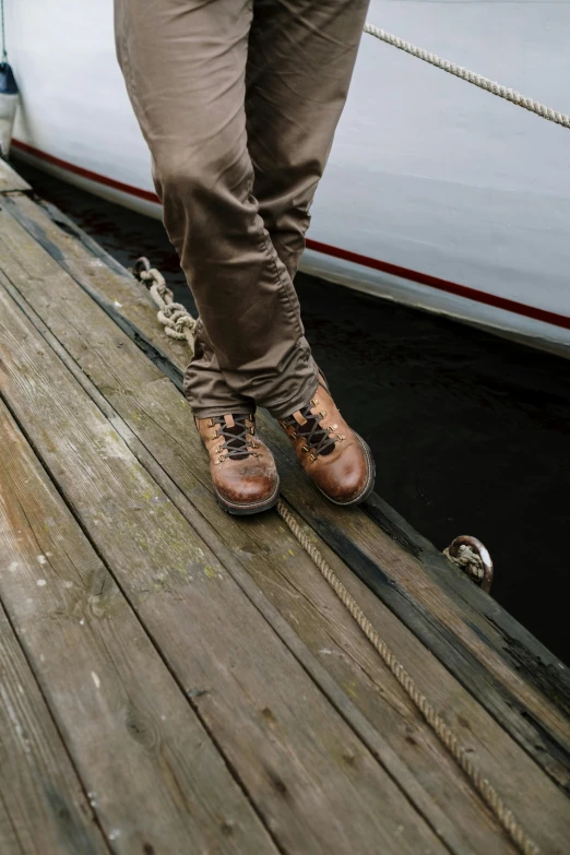 a man standing on a dock next to a boat, by Ben Zoeller, leather boots, full body extreme closeup, caramel. rugged, print