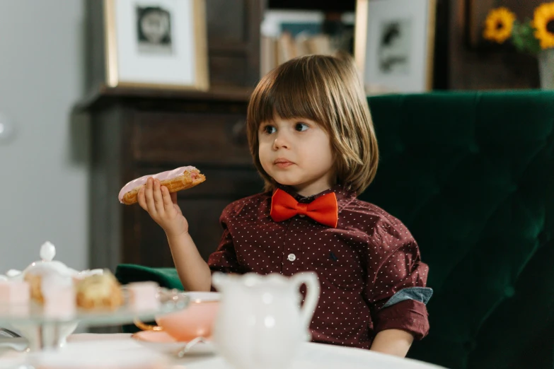 a little boy sitting at a table with a donut in his hand, by Emma Andijewska, pexels contest winner, red tailcoat, wearing a shirt with a tie, sausages, bow tie