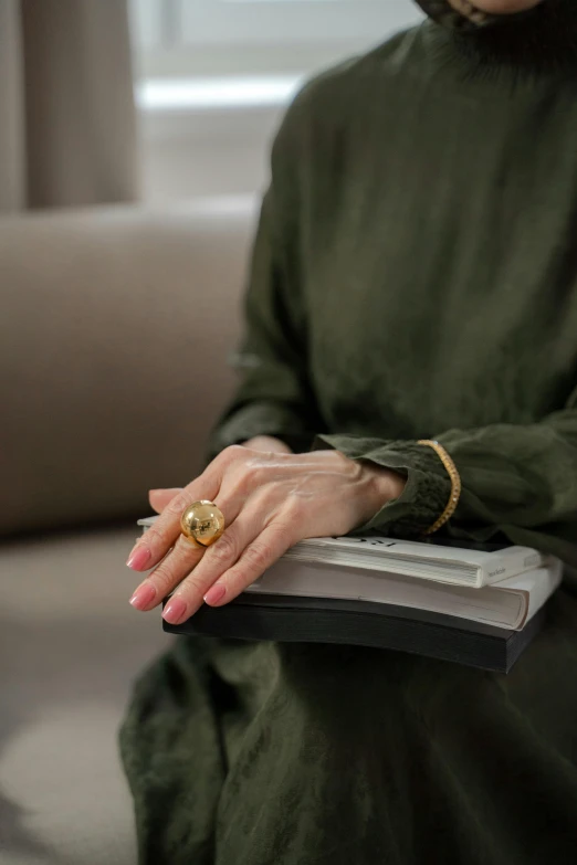 a woman sitting on a couch holding a book, by Nina Hamnett, instagram, renaissance, rings, gold plated, olive, handcrafted