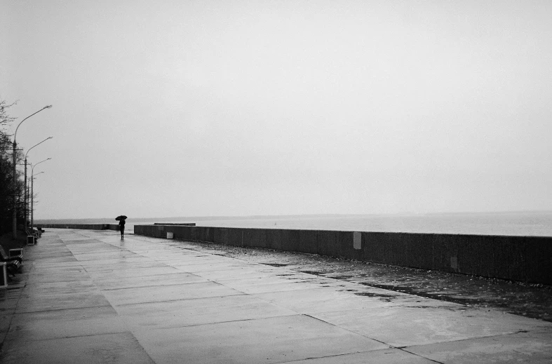 a black and white photo of a person with an umbrella, by John Hutton, minimalism, the sea seen behind the city, photographic print, wet pavement, ffffound