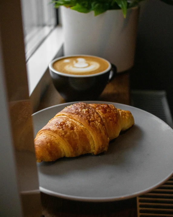 a croissant on a plate next to a cup of coffee, trending on unsplash, the window is open, lgbtq, aussie baristas, thumbnail