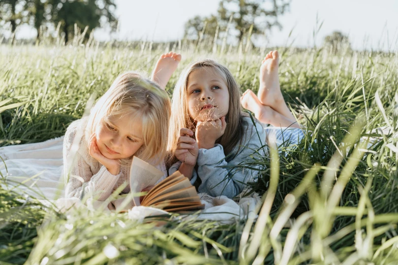 two little girls laying in the grass reading a book, inspired by Elsa Beskow, pexels contest winner, figuration libre, people on a picnic, profile image, snacks, looking towards camera