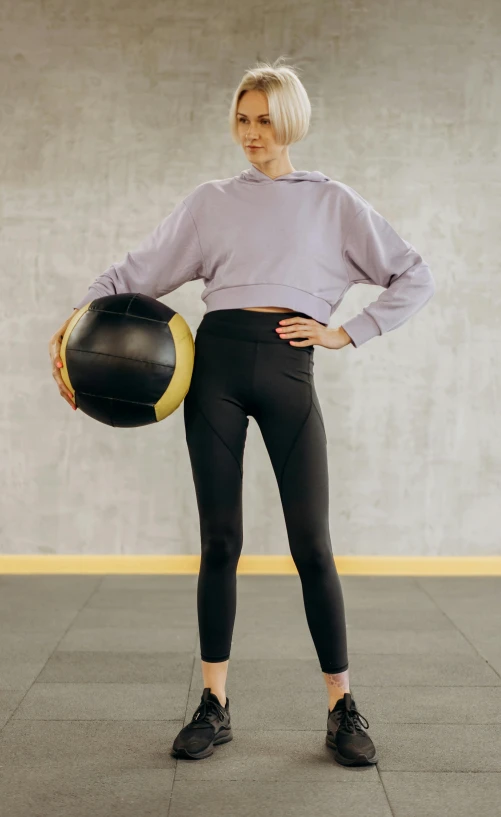 a woman standing in a gym holding a medicine ball, by Nina Hamnett, trending on pexels, realism, leggings, full body with costume, product introduction photo