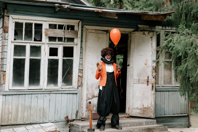 a person holding a balloon in front of a house, an album cover, by Emma Andijewska, pexels contest winner, scary clown, black and orange, black man, full body with costume