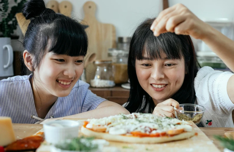 a couple of women sitting at a table with a pizza, inspired by Yukimasa Ida, trending on pexels, mingei, cooking it up, oyasumi punpun, smiling playfully, middle close up composition