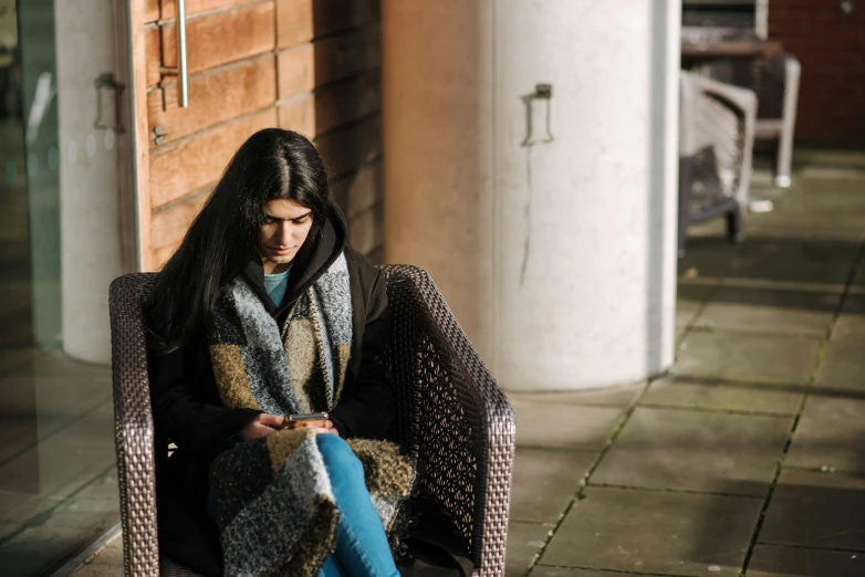 a woman sitting in a chair looking at her cell phone, by Helen Stevenson, unsplash, winter sun, indian girl with brown skin, patterned scarf, student