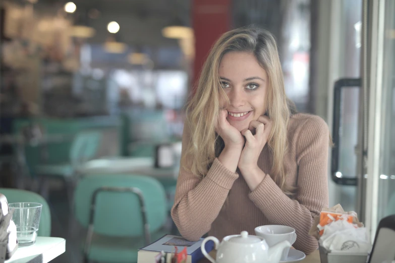 a woman sitting at a table with a cup of coffee, cheeky smile, a girl with blonde hair, multiple stories, profile image