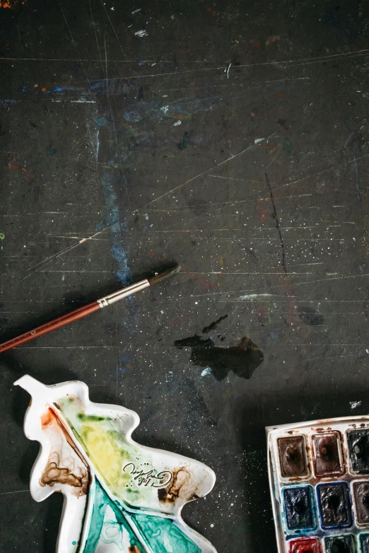 a piece of art sitting on top of a table, holding a paintbrush, miscellaneous objects, dark paint, water color