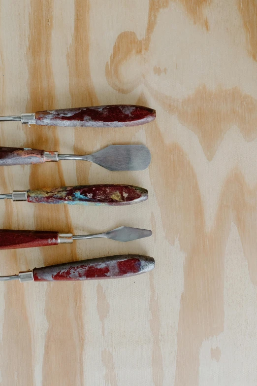 a group of knives sitting on top of a wooden table, inspired by Kyffin Williams, process art, dull red flaking paint, manuka, holding a paintbrush, detailed product image