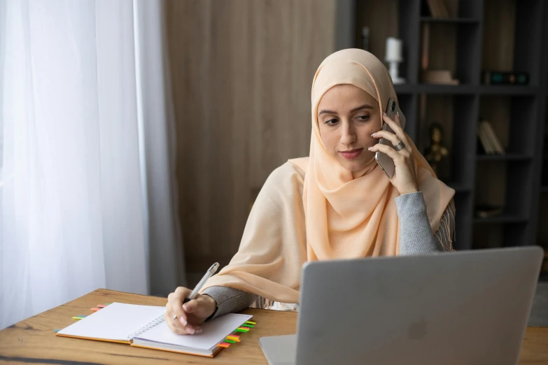 a woman sitting at a table in front of a laptop talking on a cell phone, inspired by Maryam Hashemi, hurufiyya, school curriculum expert, hijab, thumbnail, malaysian