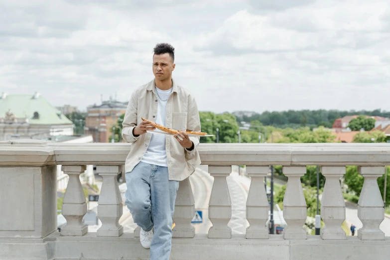 a man sitting on a ledge eating a piece of pizza, by Grytė Pintukaitė, holding a baguette, looking away from the camera, people with mohawks, promo image