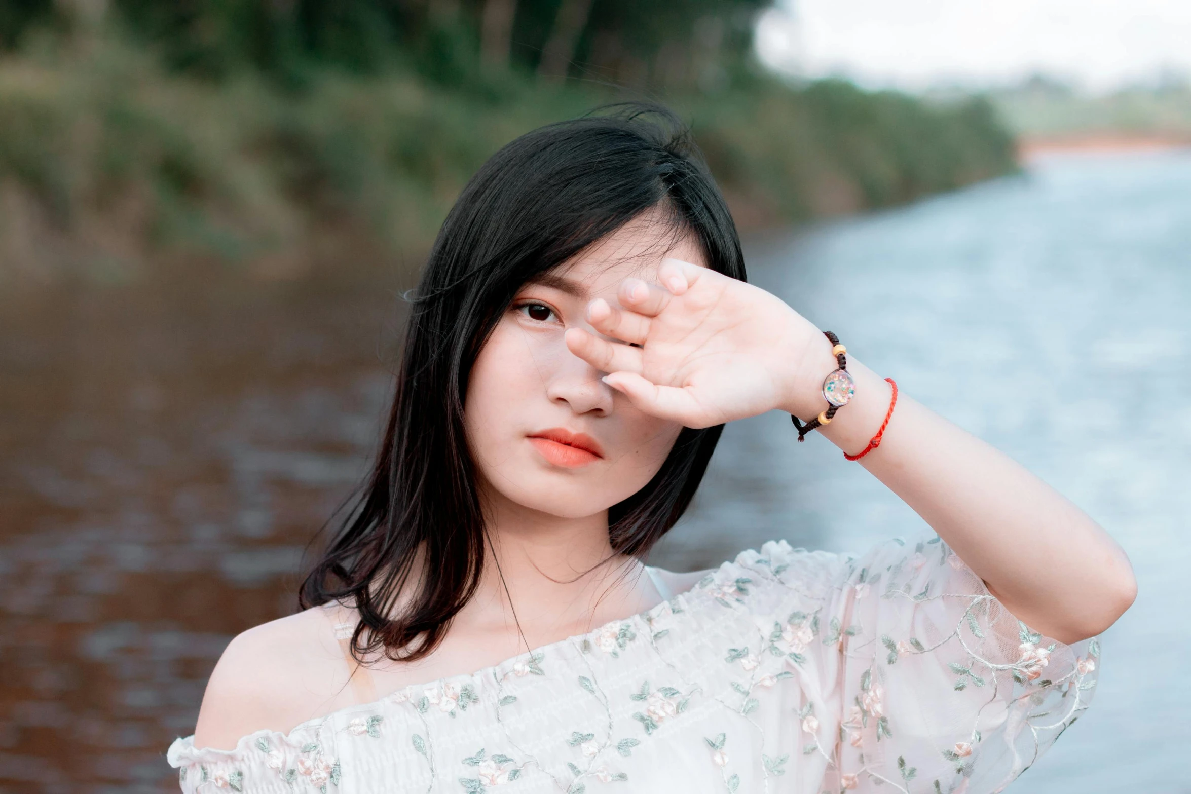 a woman standing next to a body of water, inspired by Itō Shinsui, trending on pexels, aestheticism, young adorable korean face, hands shielding face, vietnamese woman, wearing a patch over one eye