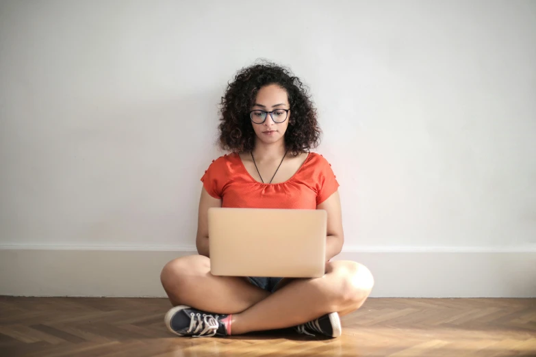 a woman sitting on the floor with a laptop, by Carey Morris, pexels, indian girl with brown skin, nerd, wearing an orange t-shirt, it specialist