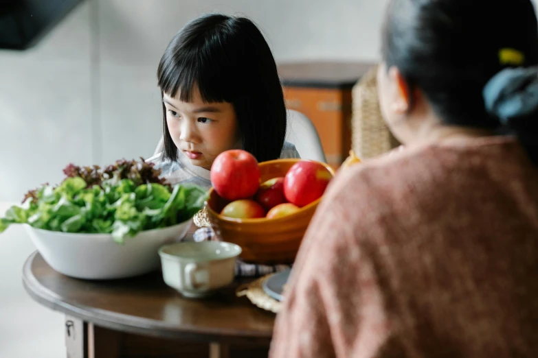 a little girl sitting at a table with a bowl of fruit and vegetables, looking at each other mindlessly, an asian woman, profile image, family friendly