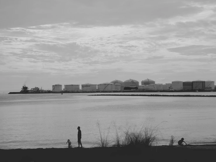 a man standing on top of a beach next to a body of water, a black and white photo, minimalism, chemical plant, long beach background, silhouettes of people, lonely family