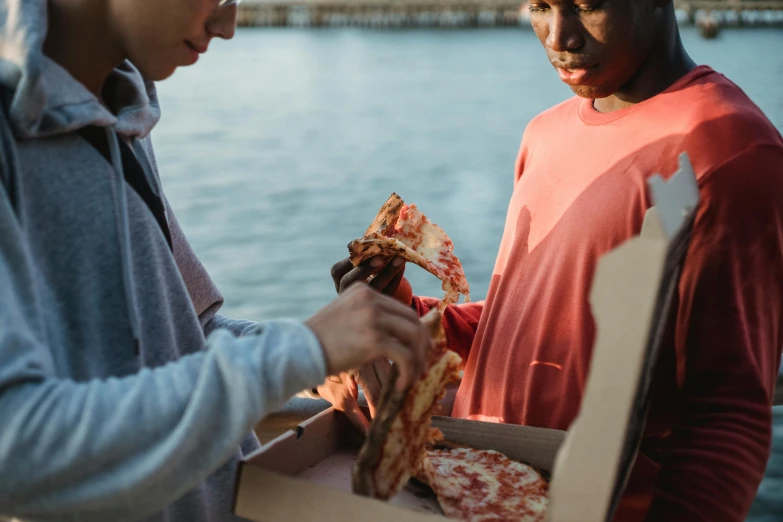 two people standing next to each other eating pizza, by Niko Henrichon, pexels contest winner, hurufiyya, delivering parsel box, at the waterside, lachlan bailey, dark-skinned