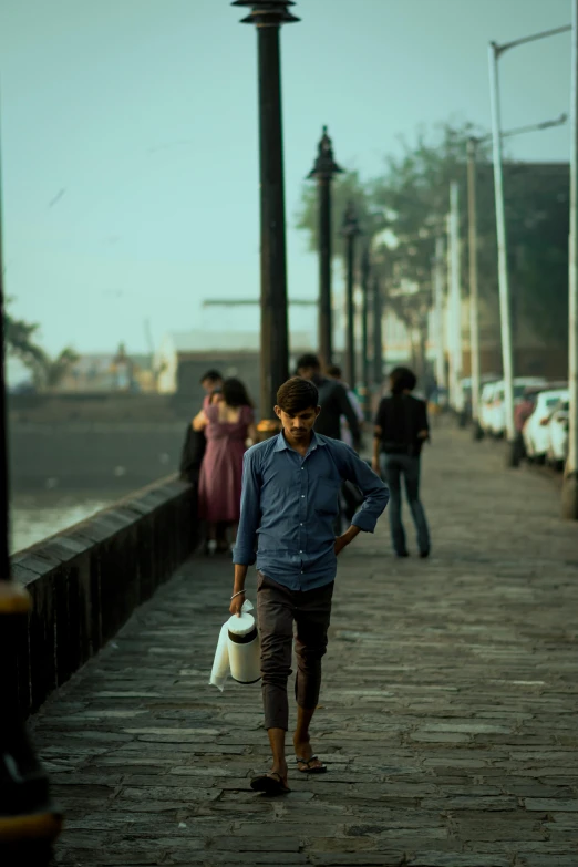 a group of people walking down a sidewalk next to a body of water, an album cover, inspired by Steve McCurry, pexels contest winner, hyperrealism, streets of mumbai, full body potrait holding bottle, 1960s color photograph, humid evening