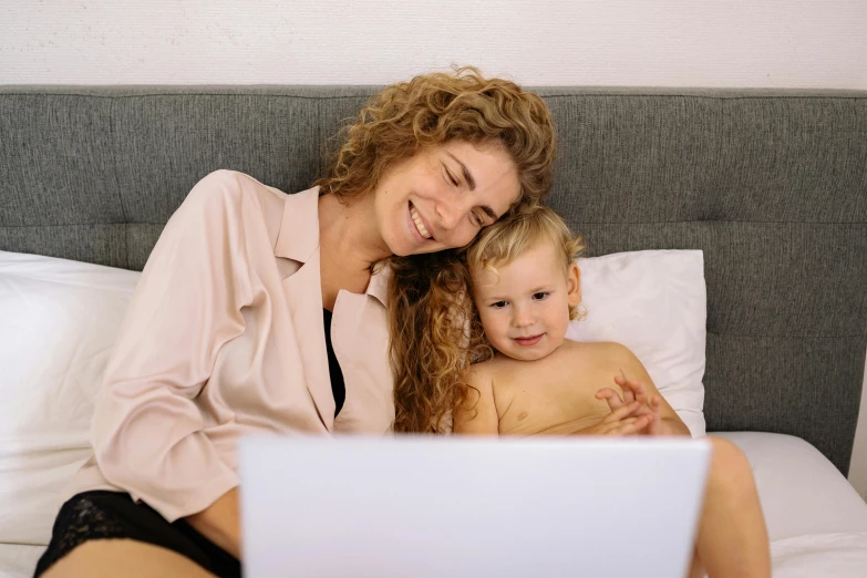 a woman sitting on top of a bed next to a baby, a cartoon, pexels, with a laptop on his lap, brown, a blond, close up to the screen