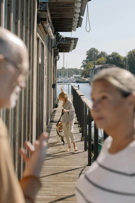 a couple of people standing on a dock next to a body of water, a picture, by Jesper Myrfors, visual art, older woman, footbridges between houses, calmly conversing 8k, directional sunlight skewed shot