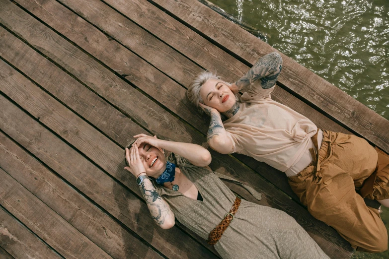 a couple of women laying on top of a wooden dock, a portrait, trending on pexels, tattooed, non-binary, james jean style, happy people