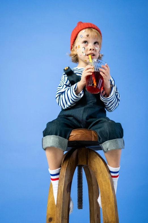 a little boy sitting on top of a wooden stool, inspired by Lucas van Leyden, pexels, pop art, with a drink, wearing a french beret, blue overalls, clown