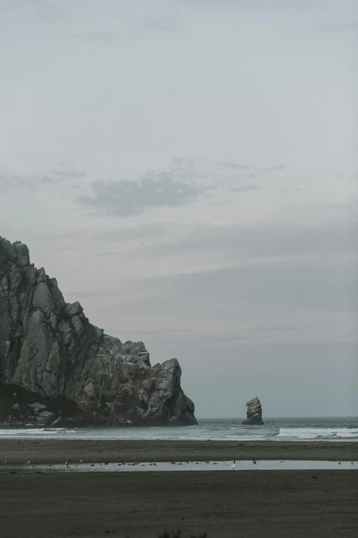 a couple of people standing on top of a sandy beach, unsplash, romanticism, big sharp rock, overcast dusk, low quality photo, ((monolith))