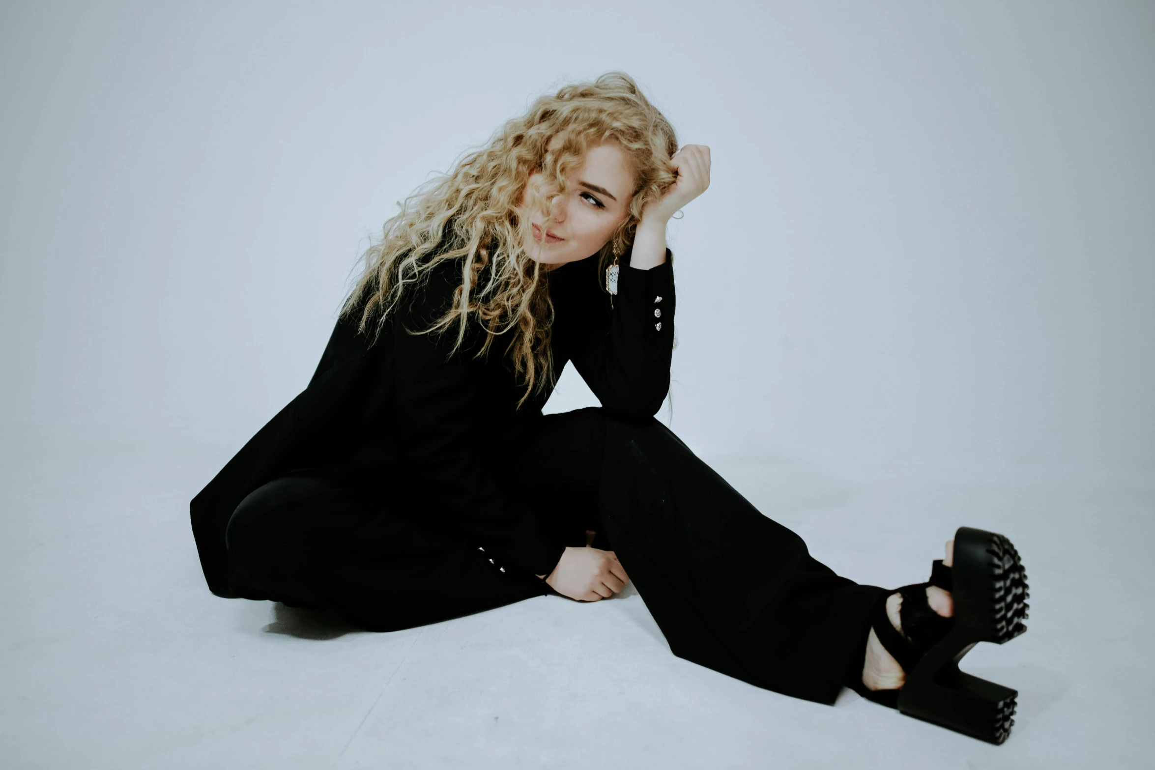 a woman sitting on the ground with her hand on her head, an album cover, pale skin curly blond hair, wearing causal black suits, on a white table, curly dirty blonde hair