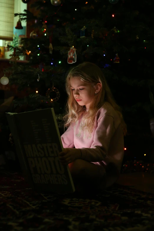 a little girl reading a book in front of a christmas tree, a picture, pexels contest winner, portrait soft low light, gif