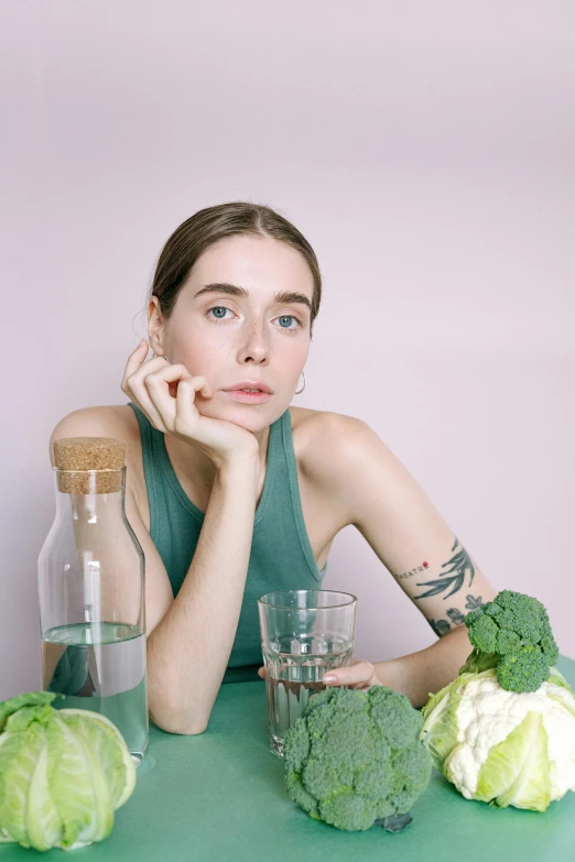 a woman sitting at a table with broccoli and water, by Jessie Alexandra Dick, trending on pexels, lilly collins, on a pale background, non binary model, looking straight forward