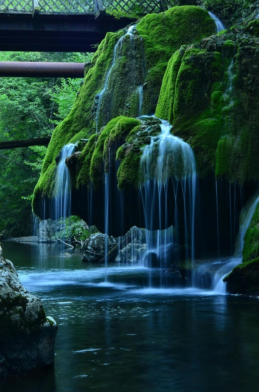 a waterfall flowing under a bridge in a forest, by Ivan Grohar, lush valley, dark green water, serenity & calm, transylvania