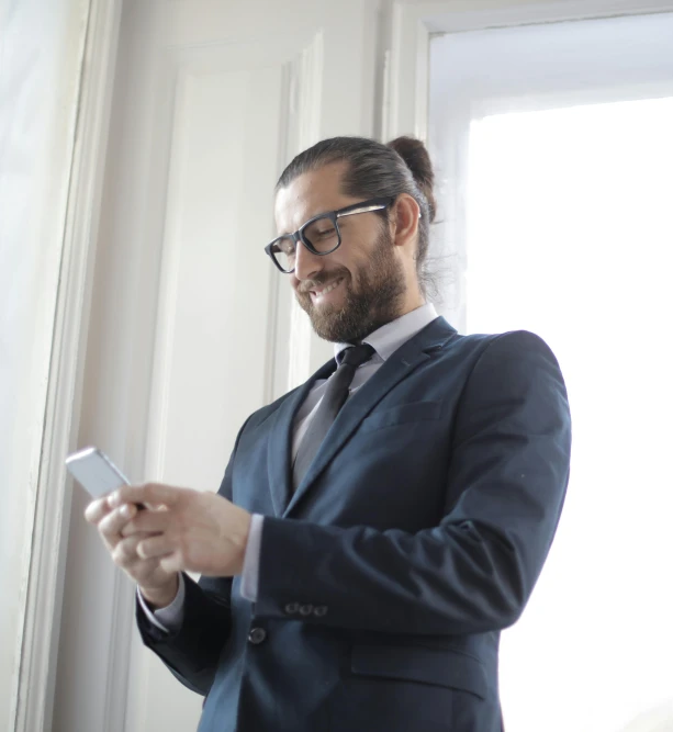 a man in a suit looking at a cell phone, ponytail and beard, standing near a window, nerdy appearance, looking happy