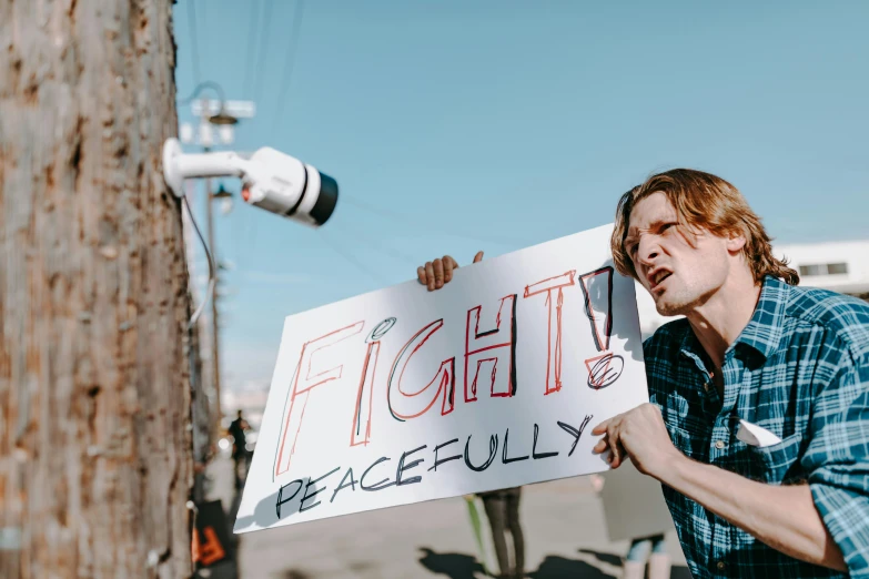 a man holding a sign in front of a pole, by Julia Pishtar, pexels, ready to fight, peace, joe keery, albuquerque