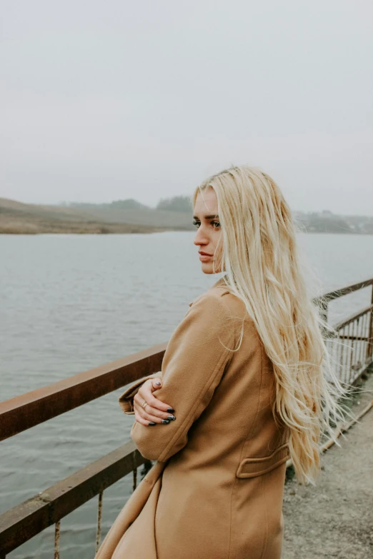 a woman standing on a pier next to a body of water, inspired by Elsa Bleda, trending on pexels, renaissance, beige hair, overcast, long golden hair, on a bridge