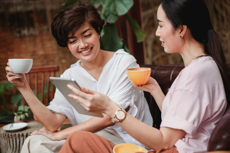 a couple of women sitting next to each other on a couch, trending on pexels, vietnamese woman, using a magical tablet, avatar image, sitting on a mocha-colored table