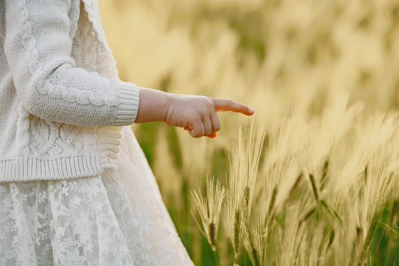 a little girl standing in a field of tall grass, inspired by Kate Greenaway, trending on pexels, symbolism, with pointing finger, wheat field, wearing a white sweater, elegantly dressed