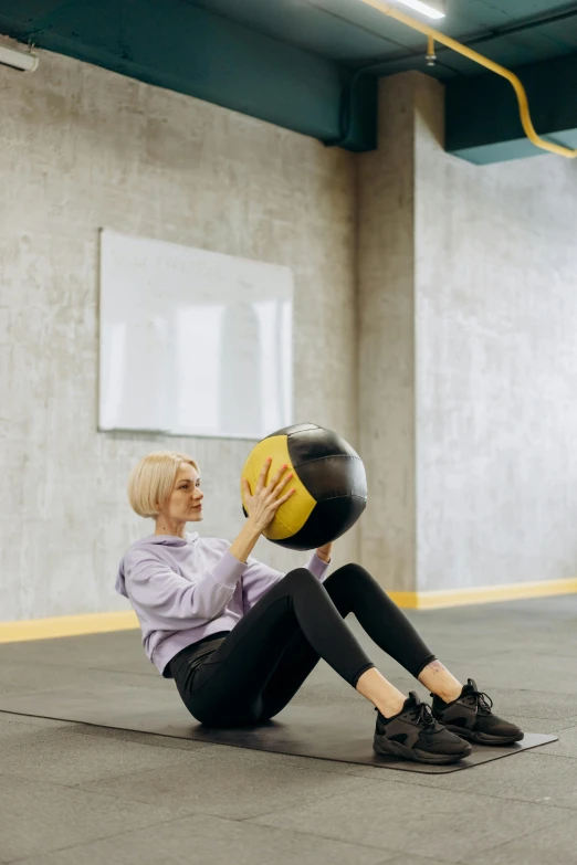 a woman sitting on the floor with a medicine ball, by Nina Hamnett, pexels contest winner, silver and yellow color scheme, athletic crossfit build, square, low ceiling