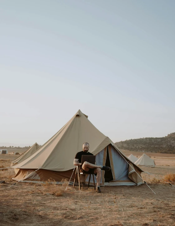 a man sitting in a chair in front of a tent, in the middle of the desert, glamping, profile image