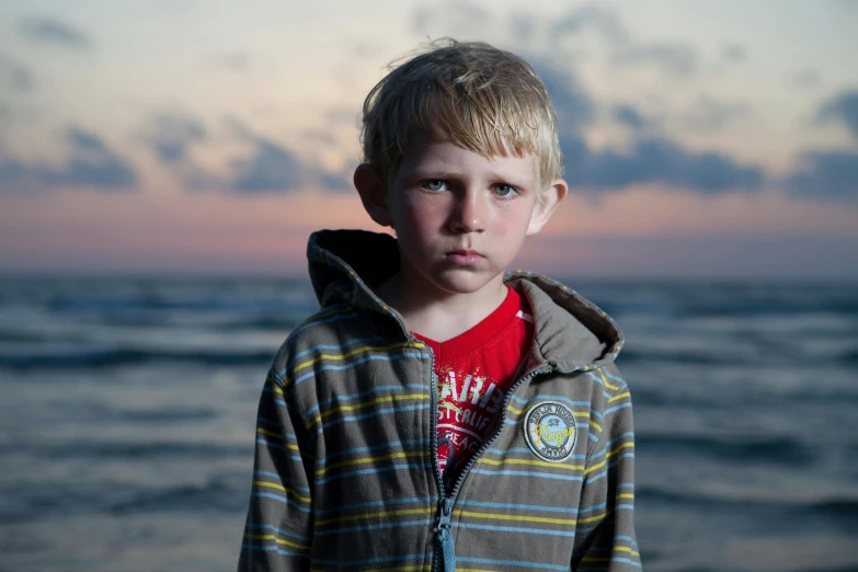 a young boy standing on top of a beach next to the ocean, an album cover, inspired by Jan Rustem, unsplash, hyperrealism, pouty face, small blond goatee, last light, concerned expression