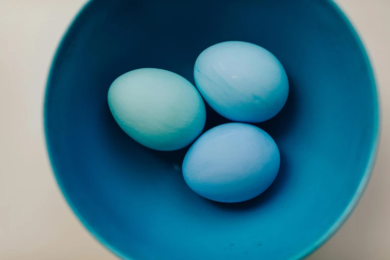 three eggs in a blue bowl on a table, by Sylvia Wishart, trending on unsplash, ((greenish blue tones)), generate multiple random colors, easter, soft blue light