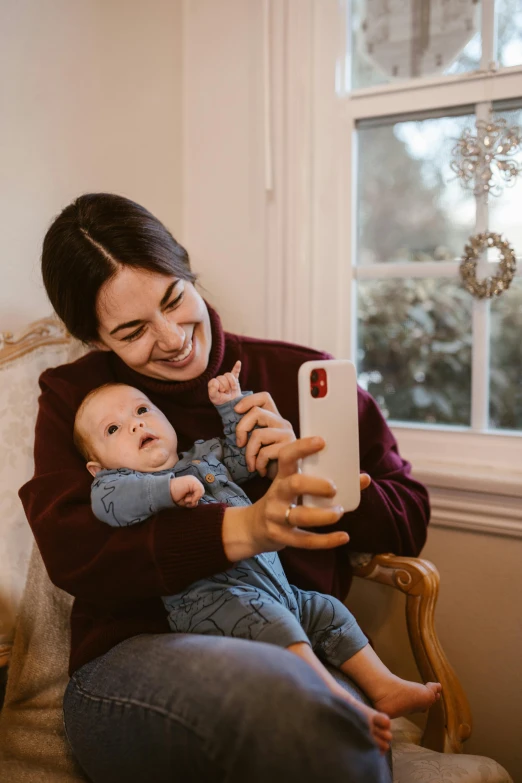 a woman sitting in a chair holding a baby, a picture, pexels contest winner, hold up smartphone, 1x, case, warm features