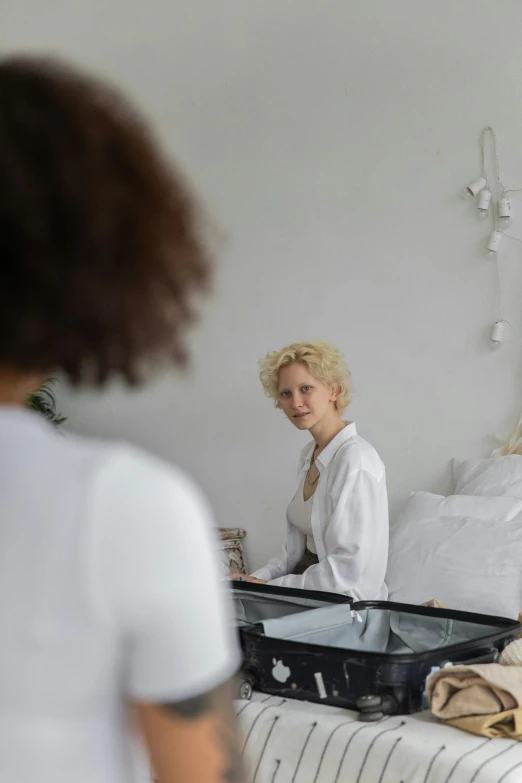 a woman sitting on a bed in front of a mirror, inspired by Vanessa Beecroft, happening, short blonde afro, wearing a white hospital gown, practical effects, looking at each other mindlessly