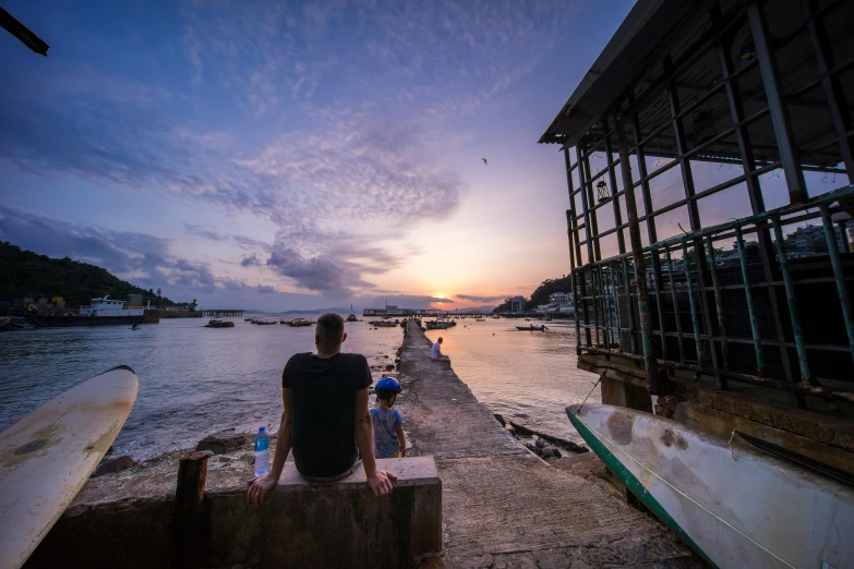 a man sitting on top of a wooden bench next to a body of water, by Sam Dillemans, pexels contest winner, happening, fishing village, late afternoon, hong kong, family friendly