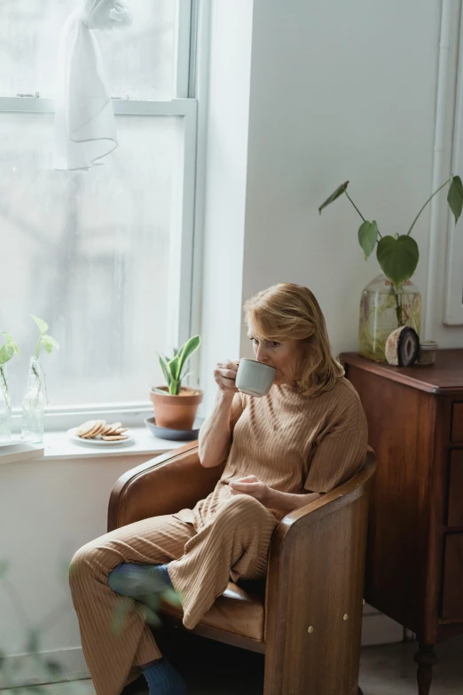a woman sitting in a chair next to a window, trending on pexels, brown sweater, wearing pajamas, daily life, julia gorokhova